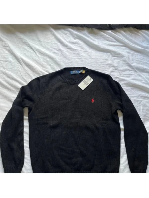 Svart stickad tröja från Polo Ralph Lauren - Klassisk svart stickad tröja från Polo Ralph Lauren med rund halsringning och den ikoniska röda logon broderad på bröstet. Perfekt till lager på lager och funkar året runt. Tröjan har långa ärmar och ribbade muddar för en clean look.