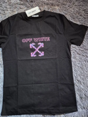 Svart Off-White t-shirt med tryck - Svart t-shirt från Off-White med färgstarkt grafiskt tryck fram och bak. Framsidan har rosa och lila Off-White-logga och pilar, medan baksidan har en stor graffiti-inspirerad figur och samma pilar. Klassisk rund hals och korta ärmar. 