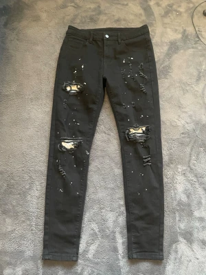 Jeans - Säljer ett par svarta jeans med slitna detaljer och färgstänk på både fram- och baksida. Byxorna har normal midja och klassisk femficksdesign. Materialet är stretchigt och bekvämt, perfekt för dig som gillar streetstil. Hör gärna av dig vid frågor!