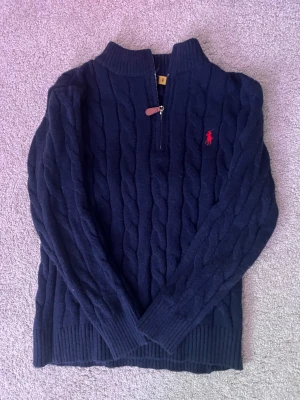 Mörkblå kabelstickad tröja Polo Ralph Lauren - Kabelstickad mörkblå tröja från Polo Ralph Lauren med röd broderad logga på bröstet. Tröjan har half zip-dragkedja, ribbade muddar och hög krage. Perfekt för kyliga dagar och ger en klassisk preppy look.