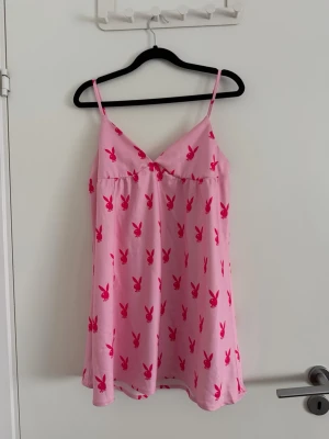 Rosa nattlinne med playboymönster - Säljer en rosa nattlinne med tunna axelband och v-ringad hals. Klänningen har ett all-over tryck med röda playboykaniner och en liten rosett framtill. Materialet känns mjukt och svalt, perfekt för sommarens alla outfits. Använd några gånger men fortfarande i fint skick.