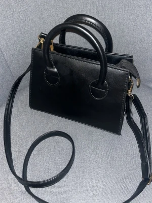 Svart stilren handväska med axelrem - Säljer en svart, stilren handväska i skinnimitation med både handtag och lång axelrem. Väskan har ett stort fack och dragkedja upptill med guldfärgade detaljer. Perfekt storlek för det viktigaste och enkel att matcha till olika outfits. Den är 21cm bred. Det får plats med massor av saker 