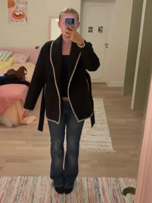 Svart omlottkappa med beige detaljer - Säljer en svart omlottkappa med beige, sicksack-broderade kanter. Jackan har bälte i midjan och är långärmad. Materialet känns som ull och modellen är loose fit, perfekt för lager på lager. Snygg och unik detalj med de kontrasterande kanterna.