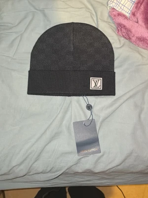 Svart Louis Vuitton mössa - Snygg svart mössa från Louis Vuitton med diskret LV-logga framtill. Klassisk form med uppvikt kant och exklusiv känsla. Tillverkad i ett mjukt material som håller dig varm och stilren under kyliga dagar. Perfekt accessoar för dig som gillar lyxiga detaljer.