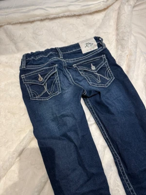 Blå bootcut jeans Gina Tricot 158 - Mörkblå jeans från Gina Tricot i storlek 158. Byxorna har tydliga vita kontrastsömmar, fickor med lock och knapp bak samt klassisk femficksmodell. Bootcut-modell med utsvängda ben och normal passform. Tillverkade i mjukt jeansmaterial.