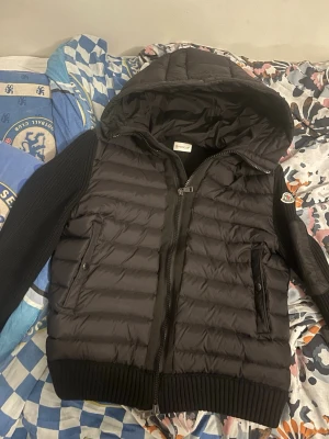 Svart Moncler cardigan med huva - Säljer en svart cardigan med storlek M men skulle nog säga att den passar S också, från Moncler med stickade ärmar och stor huva. Jackan har dragkedja framtill, två fickor med knappstängning och en extra ficka på ärmen. Klassisk helt äkta Moncler-logga på armen och ribbade muddar vid ärmslut och nederkant.