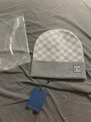 Grå rutig Louis Vuitton mössa - Snygg grå mössa från Louis Vuitton med klassiskt rutigt mönster och vikt kant. Diskret LV-logga framtill och exklusiv känsla. Perfekt accessoar för dig som vill ha något stilrent och lyxigt till vintern.