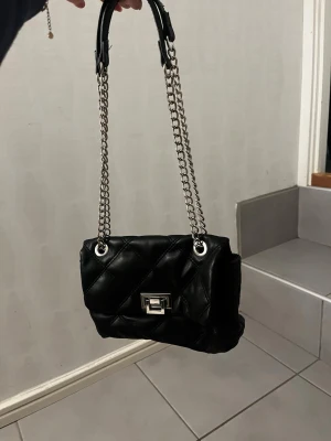 Svart quiltad axelväska med kedja - Säljer en svart quiltad axelväska med silverfärgad kedjerem och snyggt metallspänne framtill. Den har en liten defekt på spännet men annars fint skick! Bandet går att justera!