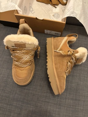 UGG lowmel - Säljer ett par snygga UGG sneakers i beige med fluffigt fårskinnsfoder och grova, mönstrade snören. Ovandelen är i mocka och textil med robust sula och rund tå. Perfekta för dig som vill ha både stil och komfort under höst och vinter.