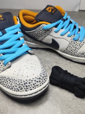 Nike SB Dunk Low Electric Safari med box och extra snören - Unika Nike SB Dunk Low med safari-mönster i grått och svart, blå snören och orange innerfoder. Skorna har svart swoosh, vita och svarta detaljer samt svart yttersula med orange Nike SB-logga. Materialmix av mocka och skinn. Extra svarta skosnören medföljer.