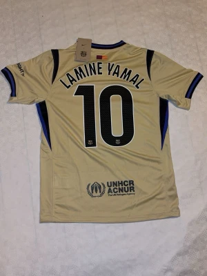 FC Barcelona Tröja LAMINE YAMAL#10 - Bilderna visar en fotbollströja från FC Barcelona med Lamine Yamal och nummer 10. Tröjan är beige med svarta och blå detaljer, Nike-märke och sponsorloggor.