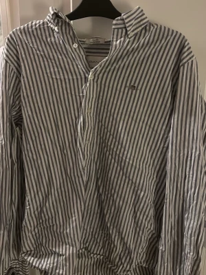 Randig skjorta från Gant Slim Fit - Säljer en klassisk vit och blå randig skjorta från Gant. Skjortan har knappar framtill, button down-krage och broderad logga på bröstet. Tillverkad i bomull med stretch för extra komfort.