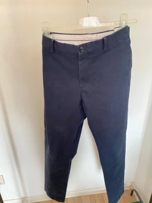 Mörkblå slim fit chinos från Riley, Storlek 50 - Snygga mörkblå chinos från Riley i slim fit-modell. Byxorna har klassisk design med bälteshällor och  bakfickor med knapp. Tillverkade i mjuk bomull med lite stretch för extra komfort. Perfekta för en clean och stilren look.