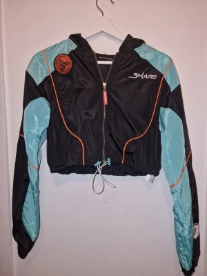 Svart och turkos vindjacka  - Cool croppad vindjacka med stor huva, broderad patch med motocross-tryck och justerbar snörning i midjan. Sportig vibe, y2k eller mer hiphop/urban stil beroende på hur du matchar. Endast tvättad vid flytt. 