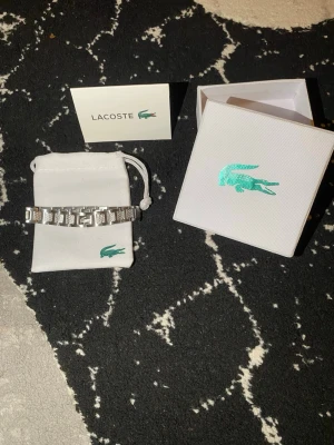 Snyggt silverfärgat armband från Lacoste - Stilrent armband från Lacoste i silverfärgad metall med länkdesign och ingraverade detaljer. Armbandet har Lacostes ikoniska krokodil på en av länkarna och kommer i original ask och tygpåse. Perfekt accessoar för att lyfta din outfit.köpte för 799 kr den är 20 cm