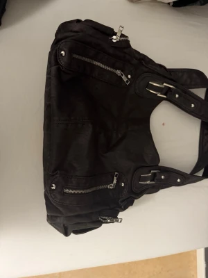 Svart axelväska med dragkedjor - Cool svart axelväska i skinnimitation med flera silvriga dragkedjor och detaljer. Väskan har breda handtag med spännen och nitar, samt flera fack både fram och bak. Perfekt för dig som vill ha en edgy och praktisk väska till vardagen.