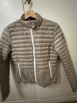 Beige pufferjacka från Colmar - Säljer en beige pufferjacka från Colmar med vita dragkedjor och diskret logga på ärmen. Jackan har quiltad design, hög krage och två fickor framtill. Insidan är ljusgrå och materialet är lätt och smidigt, perfekt för kyliga dagar.