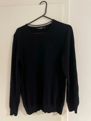 Mörkblå stickad tröja J.Lindeberg L - Snygg mörkblå stickad tröja från J.Lindeberg i storlek L. Klassisk rund halsringning och diskret broderad logga på bröstet. Perfekt till jeans eller chinos för en clean look.