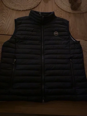 Marinblå dunväst från Colmar - Snygg svart dunväst från Colmar med quiltad design och hög krage. Västen har två fickor med dragkedja och Colmar-logga på bröstet samt en rund detalj med C på ryggen. Perfekt att slänga över en hoodie eller tröja för extra värme.      Storlek Large men passar även en längre medium.