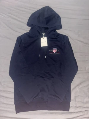 Gant Hoodie - Stilren och bekväm hoodie från GANT i klassisk mörkblå färg. Modellen har huva med dragsnören, känguruficka framtill samt ribbade muddar vid ärmslut och nederkant. GANT-logga med på bröstet ger en tidlös och sportig look. mycket fint skick och är helt oanvänd. med originaltagg kvar.