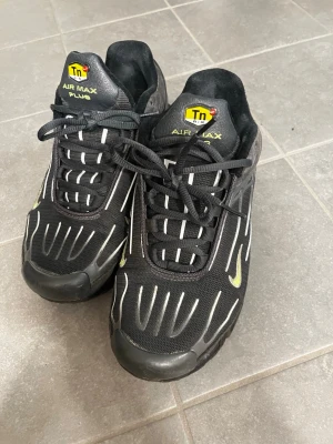 Nike Air Max Plus svarta sneakers - Säljer ett par svarta Nike Air Max Plus med vita och gula detaljer. Skorna har en mesh- och syntetöverdel, tydliga Tn-loggor på plösen och en synlig Air-sula. Snygg design med vita linjer och svart platt sula. Perfekta för dig som gillar streetwear och sportig stil.