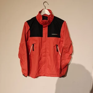 Didriksons vindjacka - Snygg röd och svart vindjacka från Didriksons1913 med huva och två dragkedjeförsedda fickor framtill. Jackan är 100% vattentät och har coola detaljer som logopatch på ärmen och kontrastpaneler. Perfekt för dig som gillar att sticka ut med färg och funktion.
