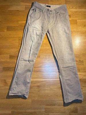 Beige chinos från EastWest - Säljer ett par klassiska beige chinos från EastWest. Byxorna har raka ben, fem fickor och snygga detaljer med metallknappar och bälteshällor. Perfekta till en avslappnad stil och tillverkade i mjuk bomull för skön känsla hela dagen.