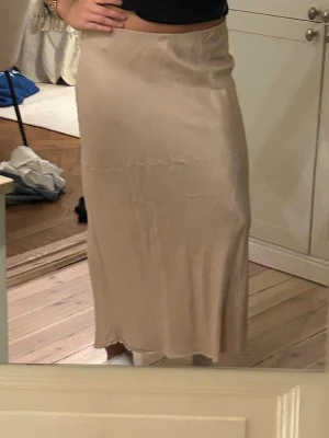 Beige satinlångkjol från Zara - Säljer en beige långkjol från Zara i glansigt satinliknande material. Kjolen har en rak och enkel siluett som sitter snyggt i midjan och faller mjukt ner mot anklarna. Perfekt att styla med en crop top eller stickad tröja. Behöver strykas