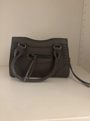 Grå handväska med dragkedja från Gina tricot - Snygg grå handväska i skinnimitation med dubbla handtag och dekorativa sömmar. Väskan har en praktisk dragkedja upptill och en mindre ficka med dragkedja framtill. Perfekt storlek för det viktigaste och enkel att matcha till olika outfits. Den är i ett väldigt bra skick. Säljer pga dne ej kommer till användning 