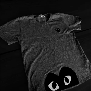 Grå t-shirt play - Säljer en grå t-shirt från Comme des Garçons Play med ikoniskt svart hjärta med ögon på bröstet och en stor svart hjärtdetalj med ögon längst ner. T-shirten har rund hals och korta ärmar, tillverkad i mjuk bomull. Perfekt för dig som gillar streetwear och coola prints.