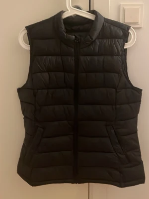 Svart quiltad dunväst från Vila - Säljer en svart quiltad dunväst från Vila med hög krage och dragkedja framtill. Västen har två fickor på sidorna och en stilren, puffig design som passar perfekt till lager-på-lager. Materialet är syntet och polyester för en lätt och bekväm känsla.