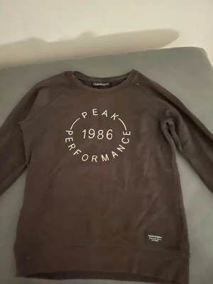 Svart Peak Performance sweatshirt - Mysig svart sweatshirt från Peak Performance med tryck 'PEAK PERFORMANCE 1986' på bröstet. Klassisk rund halsringning och ribbade muddar vid ärmslut och nederkant. Perfekt för chill dagar eller när du vill ha en enkel och snygg look.