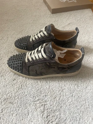 Christian Louboutin skor - Säljer ett par svarta sneakers från Louboutin med ikoniska röda sulor och coola nitar på tån. Skorna har kamouflagemönster i mocka, vita snören och detaljer i skinn. Perfekta för dig som vill sticka ut med en edgy vibe.