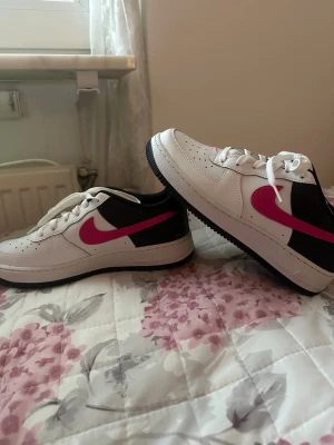 Nike Air Force 1 vit/svart/rosa( pris kan diskuteras) - Säljer ett par Nike Air Force 1 sneakers i vitt skinn med svarta detaljer och rosa swoosh. Klassisk låg modell med perforerad tå, vita skosnören och rosa innersula med Nike Air-logga. Snygg kontrast mellan färgerna och riktigt fräsch streetstil. Strl 39 , skorna är endast använda en gång.  