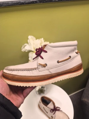 Peak Performance chukka Boots, Vit, Storlek 42 - Unika seglarskor i vitt skinn med bruna detaljer och kontrasterande vinröda snören. Skorna har klassisk mockasinsöm, beige yttersula med räfflat mönster och dekorativa läderband längs sidorna. Perfekta för dig som vill ha en stilren men ändå personlig look.