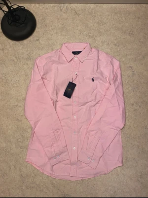 Rosa skjorta från Ralph Lauren - Säljer en klassisk rosa skjorta från Ralph Lauren med button-down krage och broderad logga på bröstet. Skjortan har långa ärmar och vita knappar framtill.
