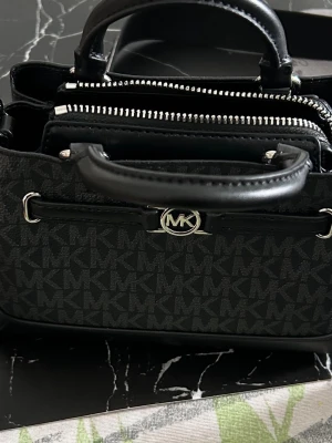 Svart handväska från Michael Kors - Snygg svart handväska från Michael Kors med klassiskt MK-mönster över hela väskan. Väskan har dubbla handtag, silverfärgade detaljer och dragkedjor samt en avtagbar axelrem. Tillverkad i skinn och syntetmaterial för en lyxig känsla.