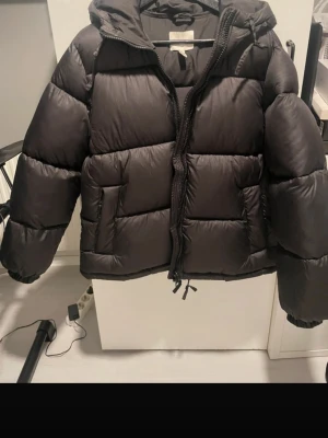 Svart pufferjacka med huva - Säljer en svart pufferjacka med huva och dragkedja framtill. Jackan är vadderad och har en boxig, lite kortare passform med snörning nertill. Perfekt för kalla dagar och riktigt skön att bära. Fickor på sidorna och elastiska muddar vid ärmsluten.