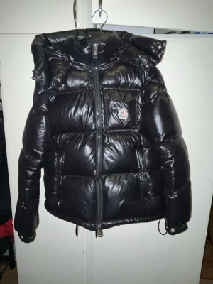 Svart Moncler Montbeliard - Säljer en svart pufferjacka från Moncler med glansig finish och stor huva. Jackan har dragkedja framtill, en bröstficka med Moncler-logga och två sidofickor. Perfekt för kalla dagar och riktigt snygg streetstil.