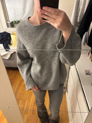 Grå stickad tröja från 157 - Mysig grå stickad tröja från 157 i storlek M. Tröjan har en relaxed passform med långa ärmar och ribbade muddar vid hals, ärmslut och nederkant. Perfekt för kyliga dagar och enkel att styla med jeans eller kjol. Den är nopprig. 