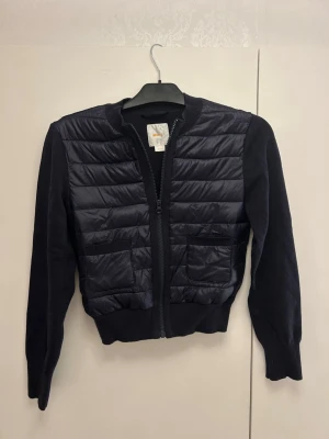 Svart quiltad jacka från Gina Tricot - Snygg svart quiltad jacka från Gina Tricot i storlek 158/164. Jackan har ribbade muddar, två fickor framtill och dragkedja. Framsidan är quiltad i blankt material medan ärmarna är stickade för en cool kontrast. Använd fåtal gånger.