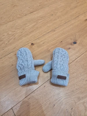 Grå vantar från Newbie 2-4år - Säljer ett par ljusgrå stickade vantar från Newbie med flätstickat mönster på ovansidan och ribbade muddar. I jättefint skick. Storlek 2-4år.