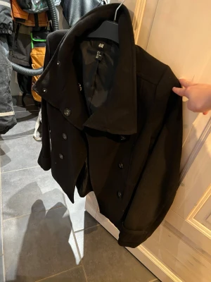 Svart skepparkavaj från H&M, strl 38 - Säljer en svart skepparkavaj från H&M i storlek 38. Jackan har dubbelknäppning framtill, bred krage och dekorativa knappar bak. Klassisk och stilren modell som är lätt att matcha till olika outfits.