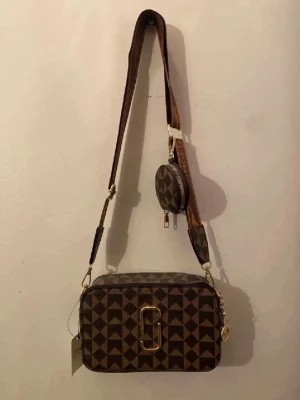 Brun axelväska med geometriskt mönster - Snygg brun axelväska med geometriskt rutigt mönster och bred axelrem med text och motiv. Väskan har guldfärgade detaljer, dubbla dragkedjor och en liten rund myntväska på remmen. Perfekt för dig som vill ha en unik accessoar med plats för det viktigaste.