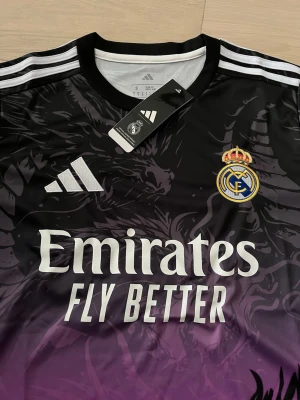 Real Madrid x Adidas draktröja svart/lila - Säljer en limited edition Real Madrid matchtröja från Adidas i svart och lila med grymt drakmotiv och vita detaljer. Tröjan har Real Madrids klubbmärke, Adidas-logga och 'Emirates Fly Better' tryckt på bröstet. Slim fit och tillverkad i polyester.
