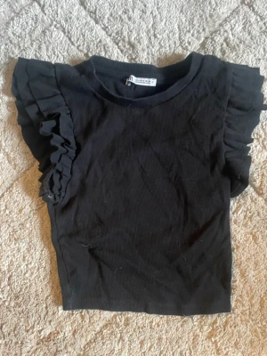 Svart volangärmad topp från Zara - Säljer en svart topp från Zara med ribbad struktur och volangdetaljer på ärmarna. Toppen har rund halsringning och är tillverkad i ett mjukt material som känns skönt mot huden. Perfekt för dig som gillar en enkel men ändå trendig look.
