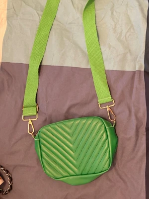 Grön axelväska med quiltad framsida - Snygg grön axelväska med bred justerbar axelrem och quiltad framsida i pilmönster. Väskan har dragkedja upptill och detaljer i guldmetall. Tillverkad i syntetmaterial och passar perfekt för att bära det viktigaste.