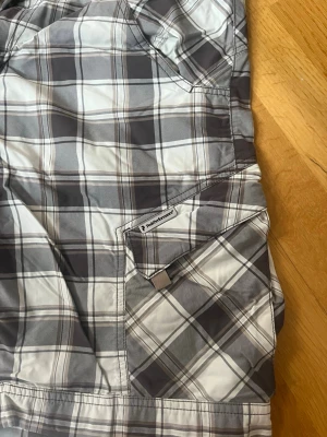 Rutiga shorts från Peak Performance - Säljer ett par rutiga shorts från Peak Performance i grå och vita toner. De har flera fickor, bälteshällor och stängs med dragkedja och knapp. Perfekta för sommardagar och riktigt sköna att bära. Klassisk cargo-look med avslappnad passform.