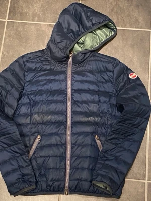 Mörkblå Colmar jacka med huva - Säljer en mörkblå jacka från Colmar med huva och två snedställda fickor med dragkedja. Jackan har quiltad design och Colmar-logga på ärmen. Perfekt för kyliga dagar och enkel att matcha med olika outfits.