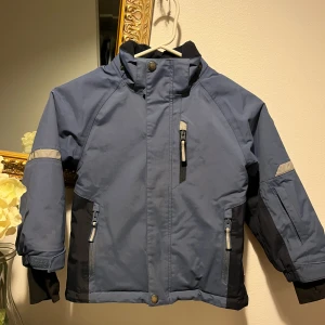 Blå vindjacka från Polarn O. Pyret - Säljer en blå och mörkblå vindjacka från Polarn O. Pyret med hög krage, reflexdetaljer och flera dragkedjeförsedda fickor. Jackan har långa ärmar med muddar och är perfekt för blåsiga dagar. Passar barn i storlek 104.